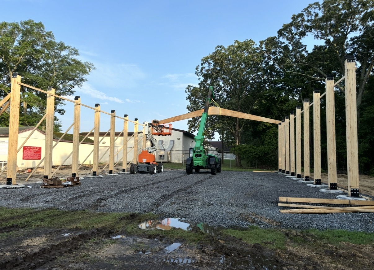 Heavy timber posts standing on the pier footings with the first glulam beam being lifted into place by a telehandler